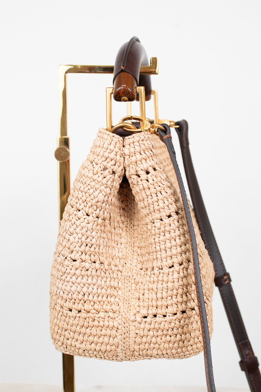 A natural beige woven straw bucket bag with a dark brown leather strap and gold-tone hardware, hanging on a gold metal display stand against a white background.
