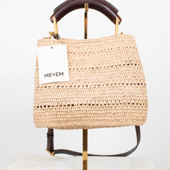 A woven straw tote bag with a dark brown leather top handle and a white ME+EM brand tag, displayed on a gold stand against a white background.