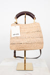 A woven straw tote bag with a dark brown leather top handle and a white ME+EM brand tag, displayed on a gold stand against a white background.