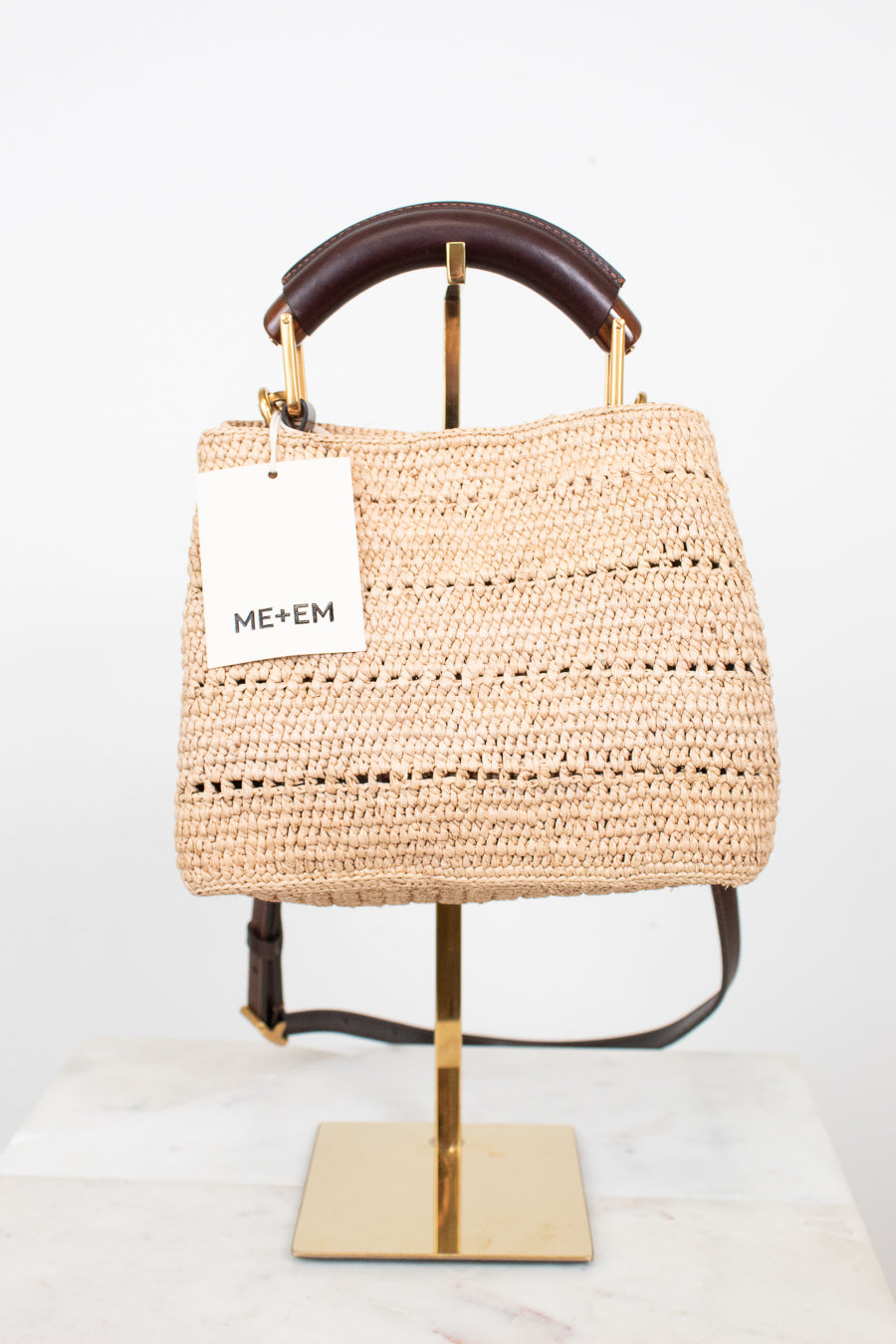 A woven straw tote bag with a dark brown leather top handle and a white ME+EM brand tag, displayed on a gold stand against a white background.