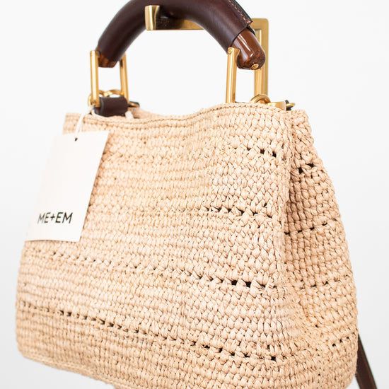 A woven straw handbag with a dark brown leather top handle and gold-tone hardware, displayed on a stand against a white background. A white tag with the brand name 'ME+EM' is attached to the side.