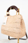 A woven straw handbag with a dark brown leather top handle and gold-tone hardware, displayed on a stand against a white background. A white tag with the brand name 'ME+EM' is attached to the side.