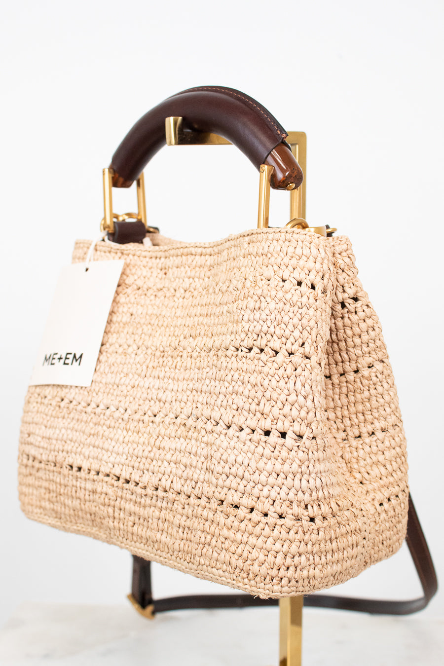 A woven straw handbag with a dark brown leather top handle and gold-tone hardware, displayed on a stand against a white background. A white tag with the brand name 'ME+EM' is attached to the side.