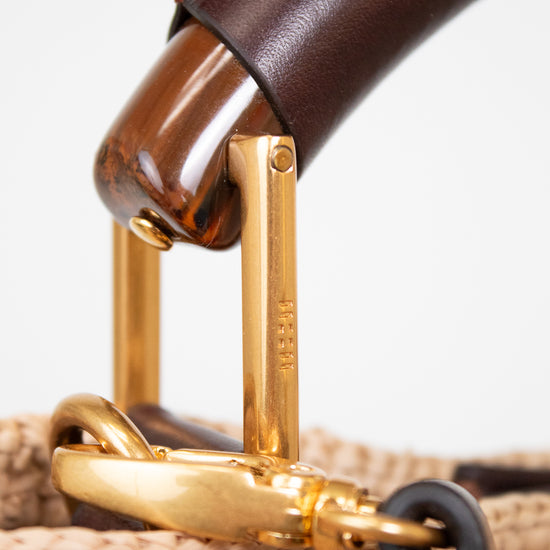 Close-up detail of a woven straw handbag handle with dark brown leather, polished wood, and gold-tone metal hardware on a white background.
