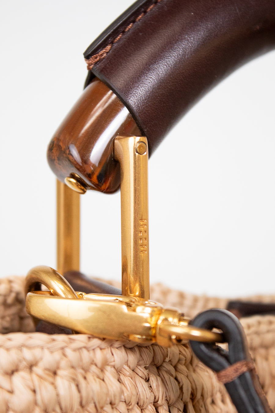 Close-up detail of a woven straw handbag handle with dark brown leather, polished wood, and gold-tone metal hardware on a white background.