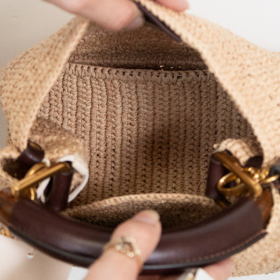 Close-up of a woven straw handbag being held open by two hands, showing the tan ribbed fabric lining and dark brown leather handles with gold-tone hardware.