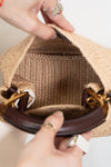 Close-up of a woven straw handbag being held open by two hands, showing the tan ribbed fabric lining and dark brown leather handles with gold-tone hardware.