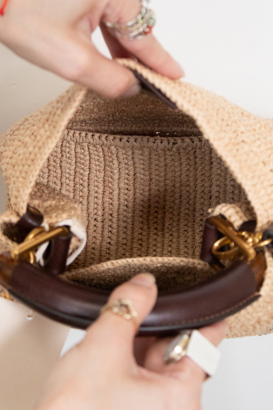 Close-up of a woven straw handbag being held open by two hands, showing the tan ribbed fabric lining and dark brown leather handles with gold-tone hardware.