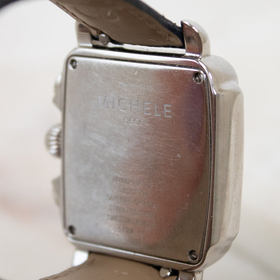 Close-up of the back of a square metal watch case with a leather strap, showing engraved text that reads 'MICHELE'.
