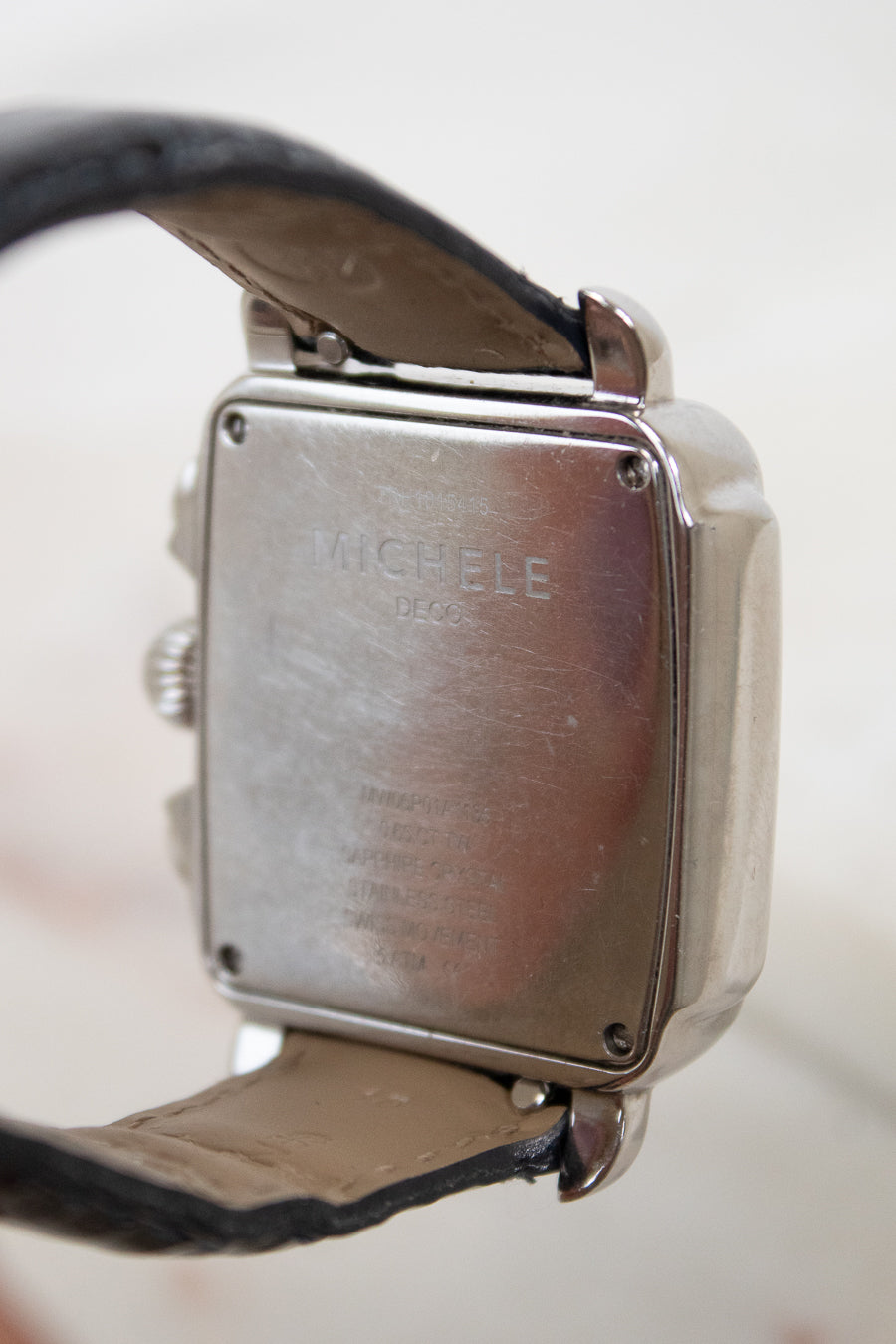 Close-up of the back of a square metal watch case with a leather strap, showing engraved text that reads 'MICHELE'.