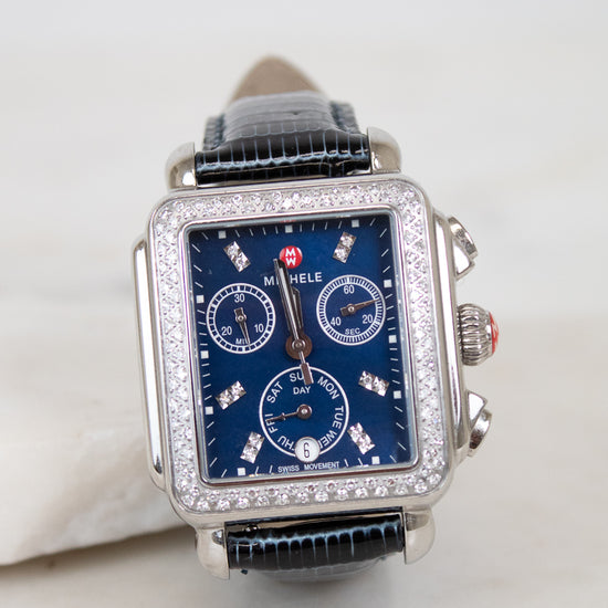 A rectangular silver-tone watch with a blue dial, diamond embellishments around the bezel and sub-dials, and a navy blue lizard leather strap, photographed on a light neutral background.