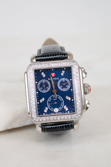 A rectangular silver-tone watch with a blue dial, diamond embellishments around the bezel and sub-dials, and a navy blue lizard leather strap, photographed on a light neutral background.