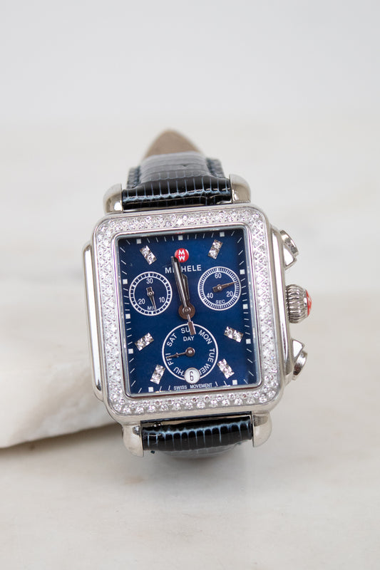 A rectangular silver-tone watch with a blue dial, diamond embellishments around the bezel and sub-dials, and a navy blue lizard leather strap, photographed on a light neutral background.