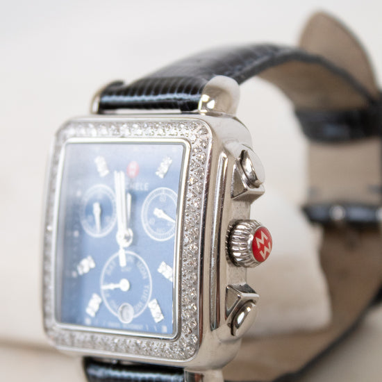 A luxury wristwatch with a silver-tone rectangular case encrusted with clear stones, a blue dial with three sub-dials, and a black leather strap.
