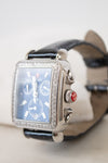 A luxury wristwatch with a silver-tone rectangular case encrusted with clear stones, a blue dial with three sub-dials, and a black leather strap.