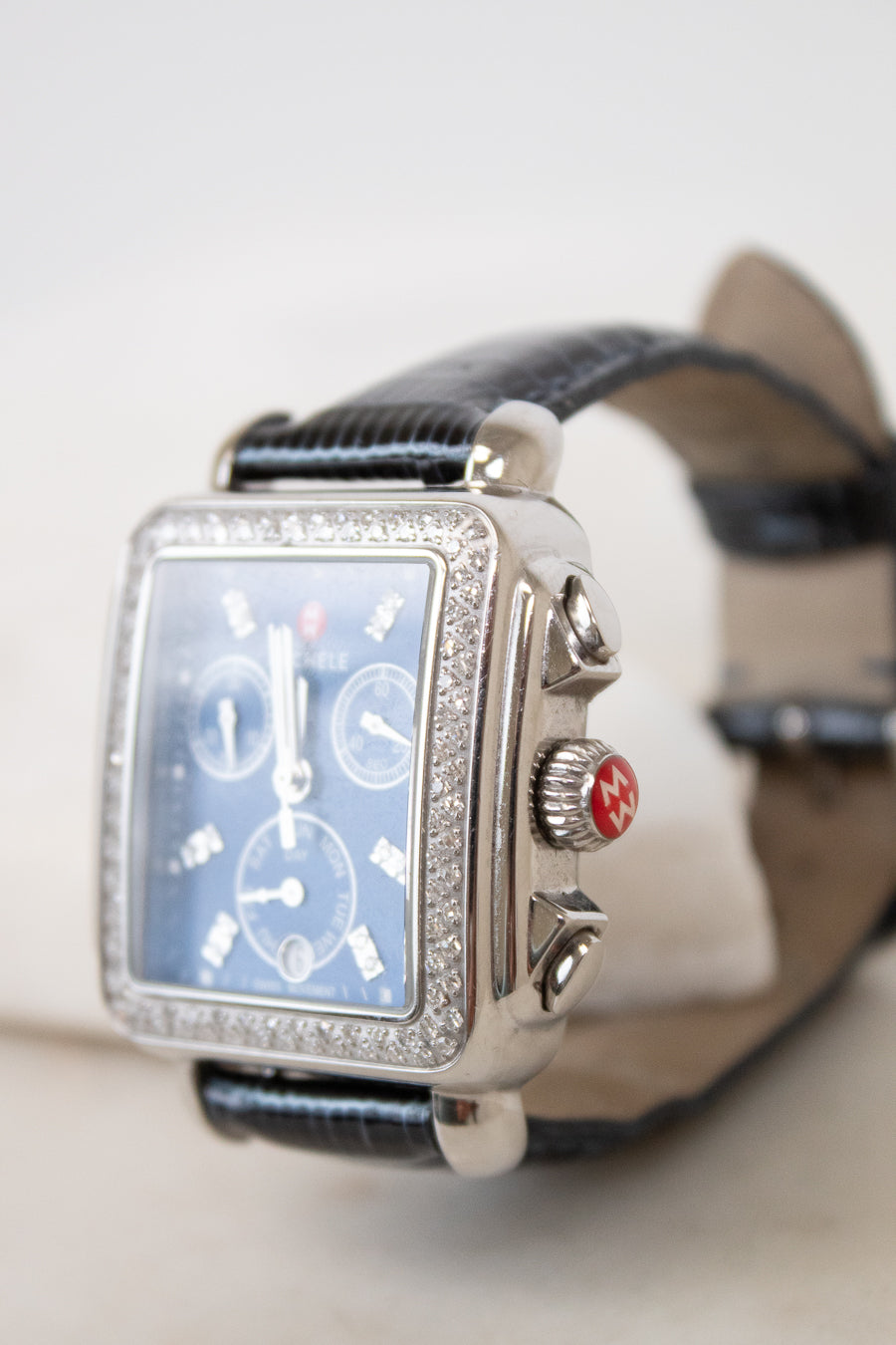 A luxury wristwatch with a silver-tone rectangular case encrusted with clear stones, a blue dial with three sub-dials, and a black leather strap.