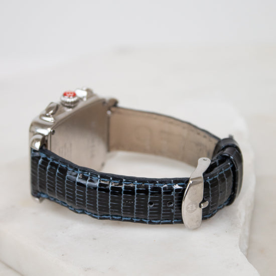 A vintage-style watch with a textured dark blue lizard-pattern strap and a silver-toned case, resting on a white stone block against a white background.