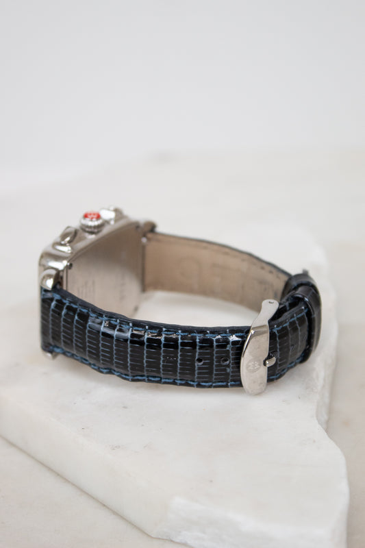 A vintage-style watch with a textured dark blue lizard-pattern strap and a silver-toned case, resting on a white stone block against a white background.