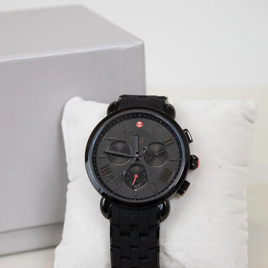 Black wristwatch with detailed face on a white cushion, gray box in the background