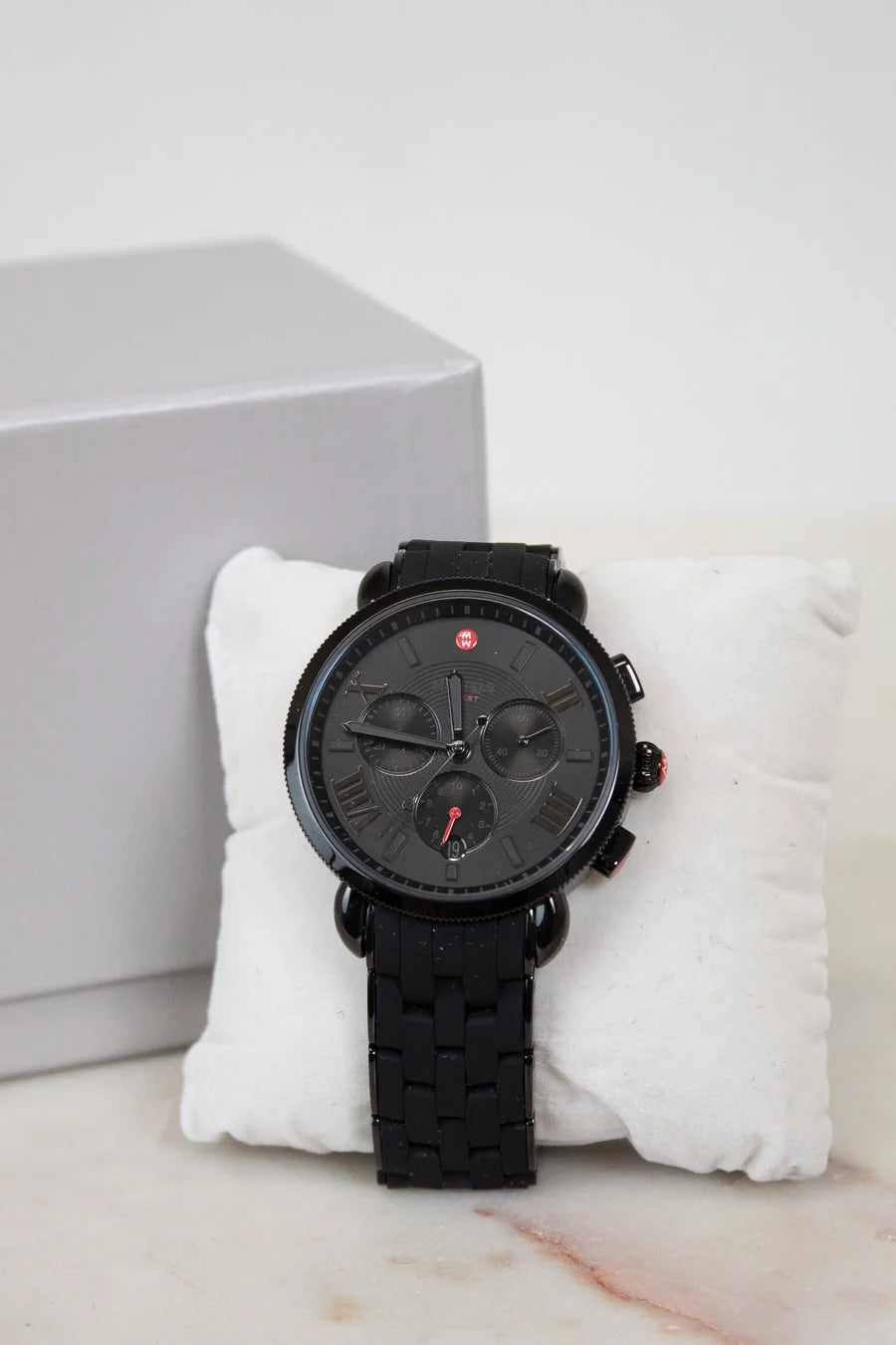 Black wristwatch with detailed face on a white cushion, gray box in the background