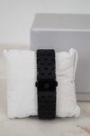 Black watch with textured strap on a white cushion against a light gray background