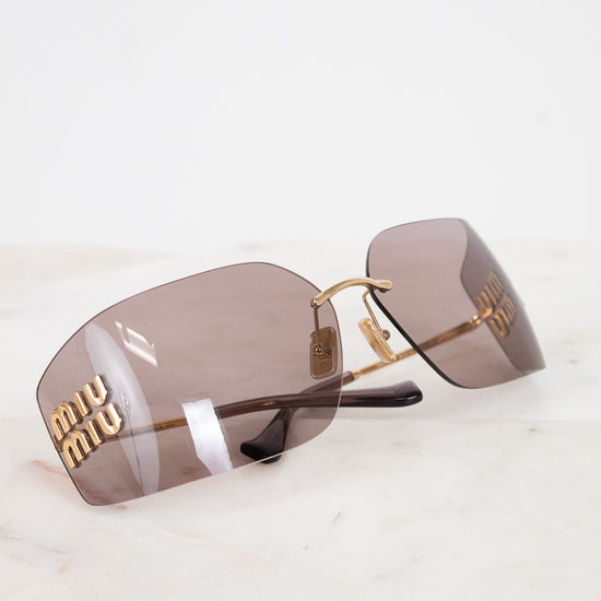 Miu Miu rimless sunglasses with gold metal frames and brown-tinted lenses, resting on a white marble surface.