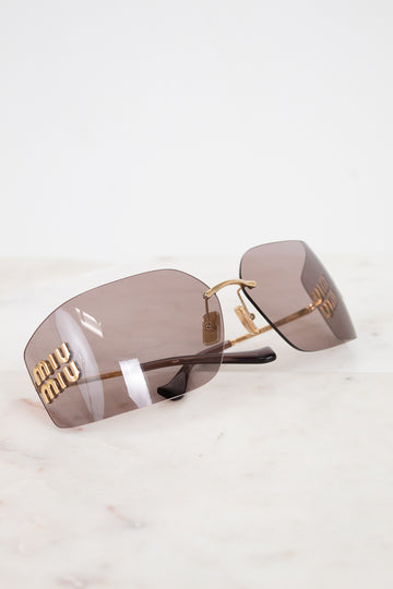Miu Miu rimless sunglasses with gold metal frames and brown-tinted lenses, resting on a white marble surface.