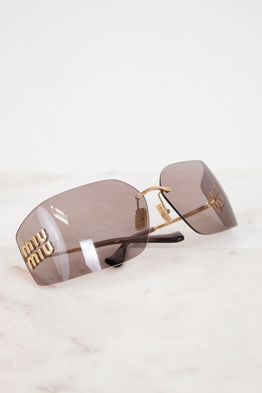Miu Miu rimless sunglasses with gold metal frames and brown-tinted lenses, resting on a white marble surface.