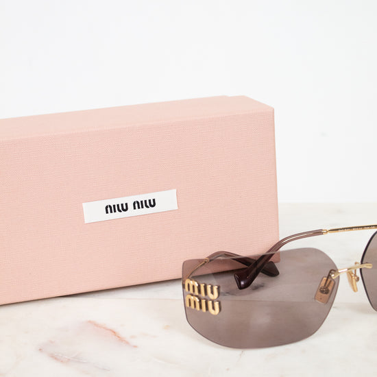 Miu Miu sunglasses with gold-tone metal frames, brown tinted lenses, and signature Miu Miu lettering on the temple, displayed next to their pink presentation box on a white marble surface.