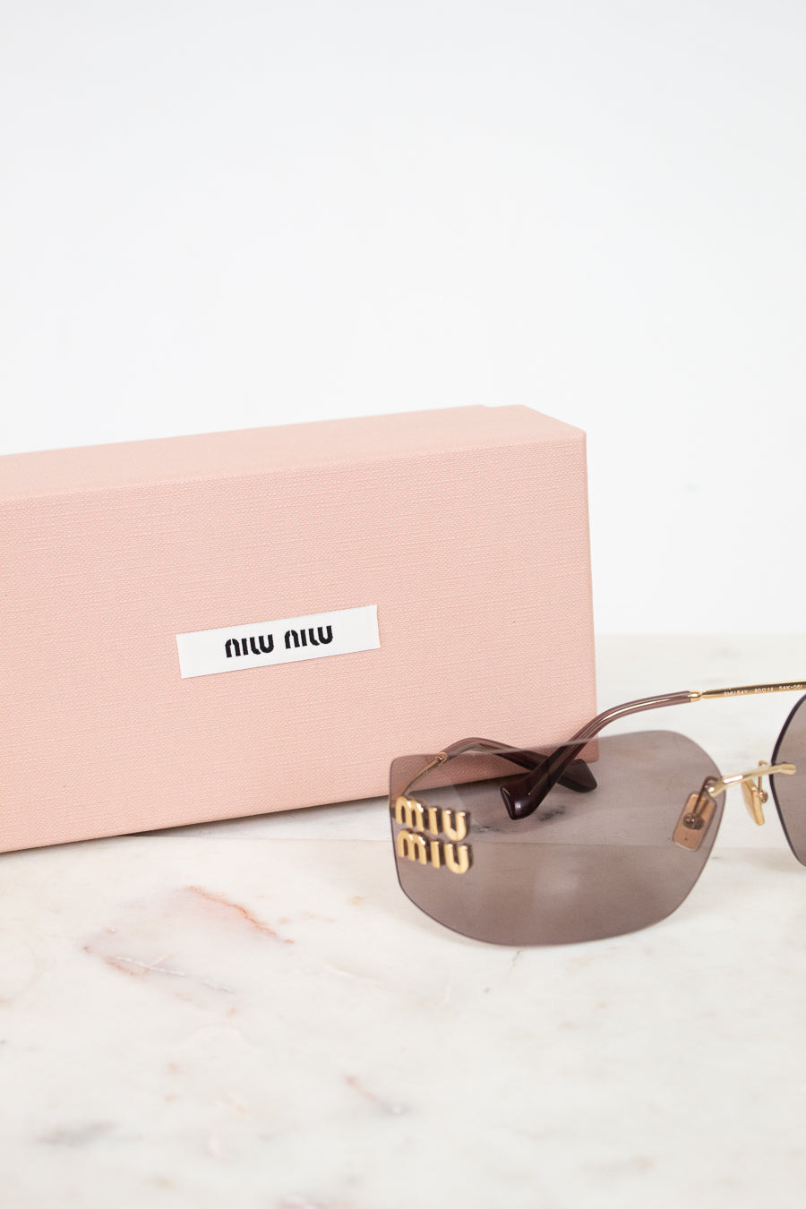 Miu Miu sunglasses with gold-tone metal frames, brown tinted lenses, and signature Miu Miu lettering on the temple, displayed next to their pink presentation box on a white marble surface.