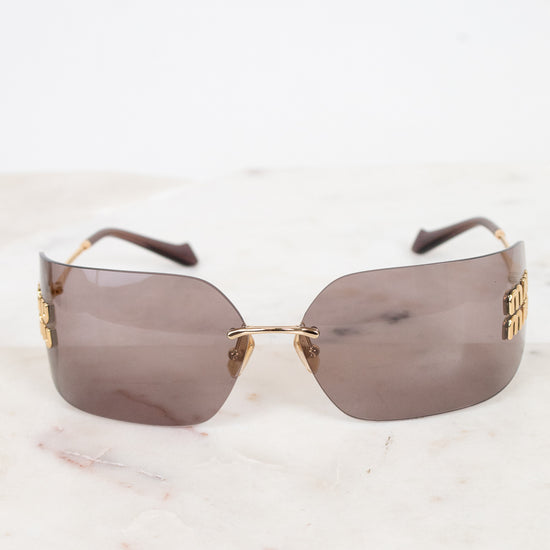 Rimless sunglasses with a gold metal logo and arm detail and tinted brown lenses, resting on a white marble surface against a white background.