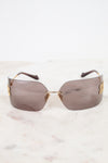 Rimless sunglasses with a gold metal logo and arm detail and tinted brown lenses, resting on a white marble surface against a white background.