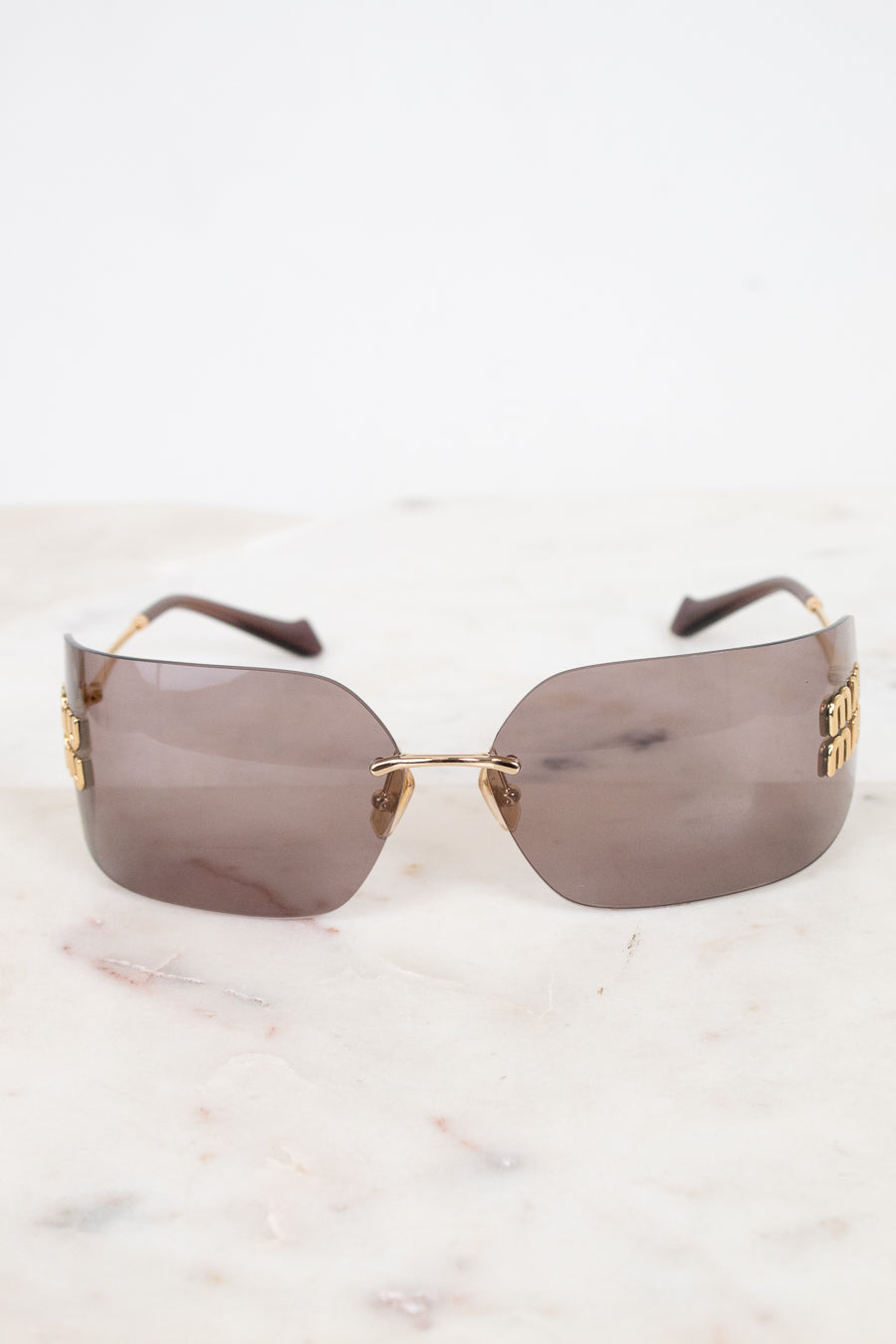 Rimless sunglasses with a gold metal logo and arm detail and tinted brown lenses, resting on a white marble surface against a white background.