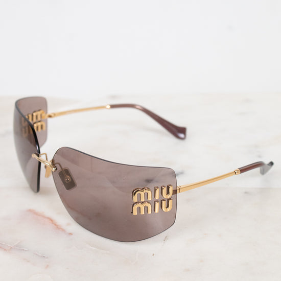Miu Miu shield-shaped sunglasses with gold metal details on arms and logo and brown-tinted lenses, resting on a white marble surface against a white background.