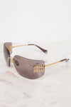 Miu Miu shield-shaped sunglasses with gold metal details on arms and logo and brown-tinted lenses, resting on a white marble surface against a white background.