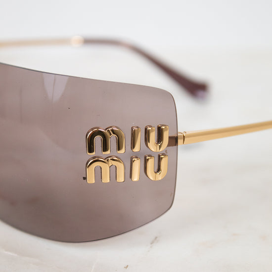 Close-up of Miu Miu shield-style sunglasses with a translucent taupe lens and gold-tone metal arms, featuring the brand name 'miu miu' in gold lettering on the side.