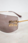 Close-up of Miu Miu shield-style sunglasses with a translucent taupe lens and gold-tone metal arms, featuring the brand name 'miu miu' in gold lettering on the side.