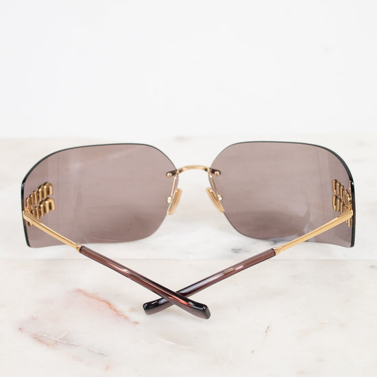 Rimless sunglasses with rectangular brown-tinted lenses, gold metal frames, and branded temples with gold Miu Miu logo details, displayed on a white marble surface.