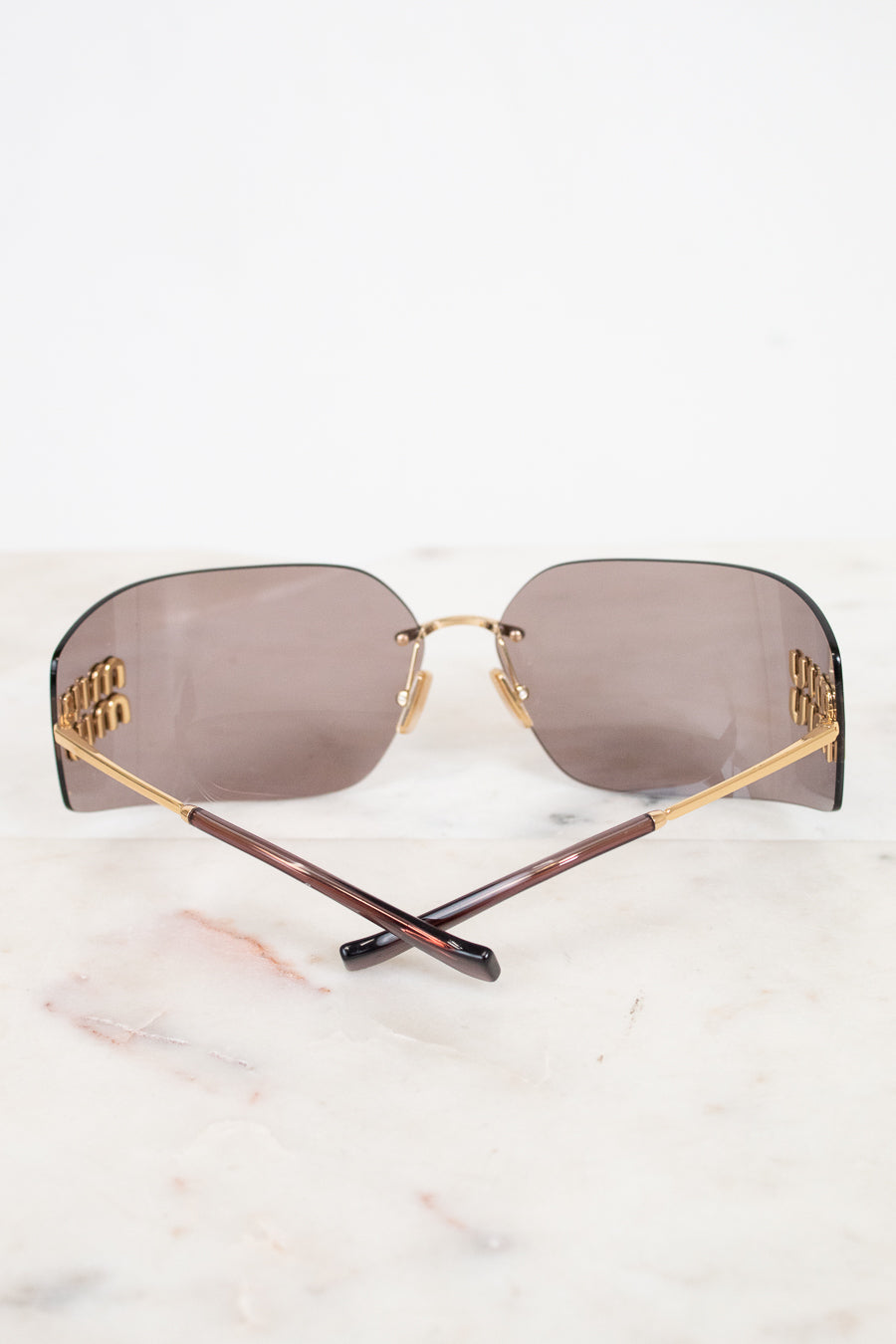 Rimless sunglasses with rectangular brown-tinted lenses, gold metal frames, and branded temples with gold Miu Miu logo details, displayed on a white marble surface.