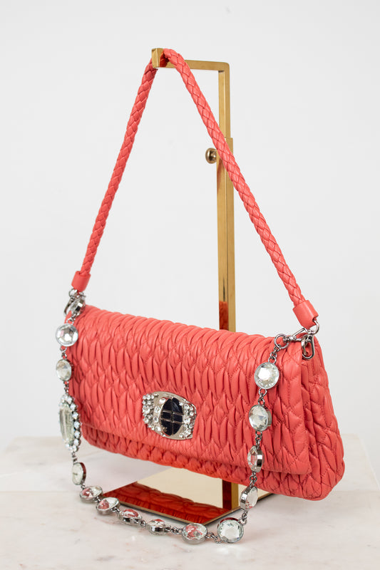 Coral handbag with decorative chain and clasp on a stand against a white background