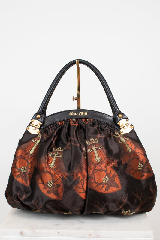 A Miu Miu handbag with black leather handles and a black satin body featuring a brown and orange pattern of keyhole silhouettes with crosses, displayed on a white surface against a white background.