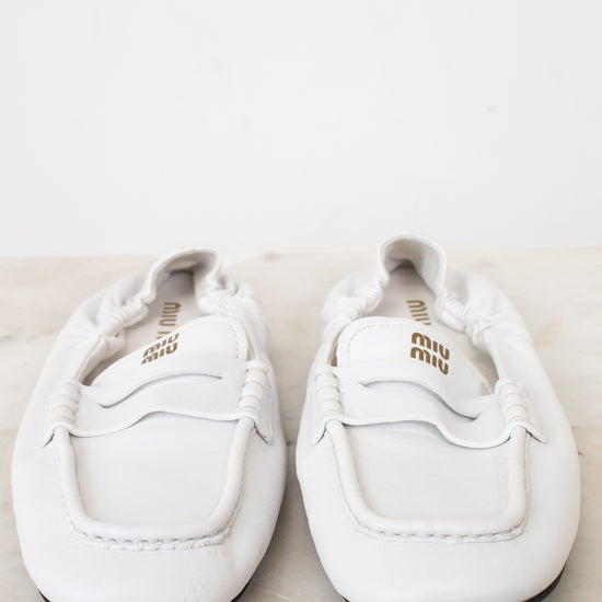 A pair of white leather Miu Miu loafers with gold branding, displayed on a light marble surface against a white background.