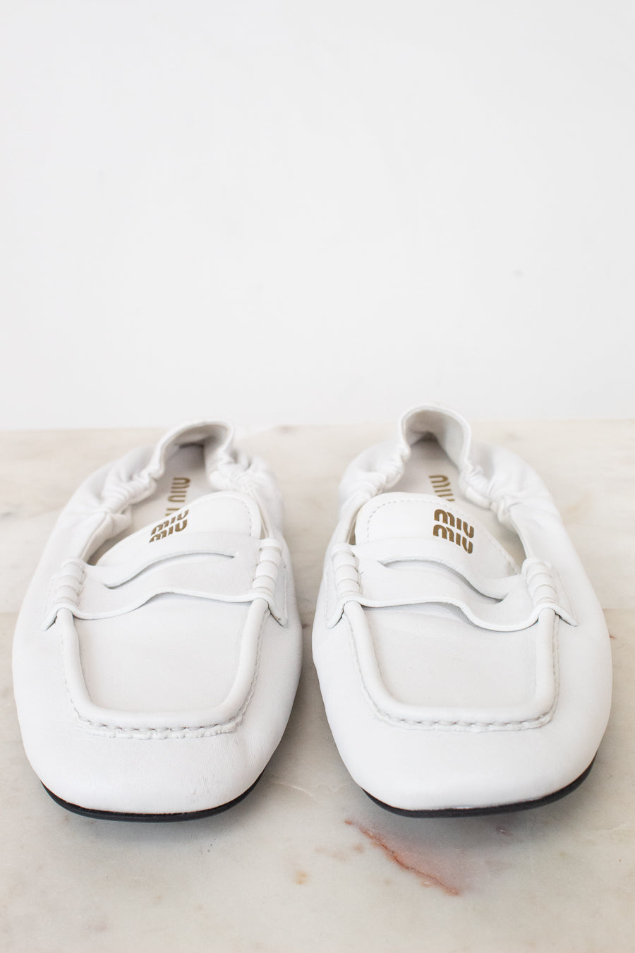 A pair of white leather Miu Miu loafers with gold branding, displayed on a light marble surface against a white background.