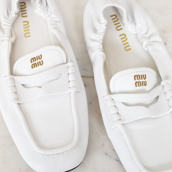 A pair of white Miu Miu leather penny loafers with gold branding, displayed on a light marble surface.
