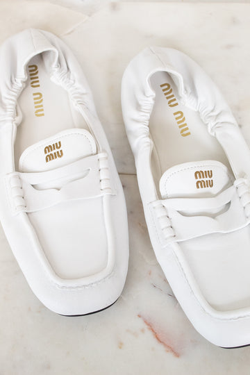 A pair of white Miu Miu leather penny loafers with gold branding, displayed on a light marble surface.