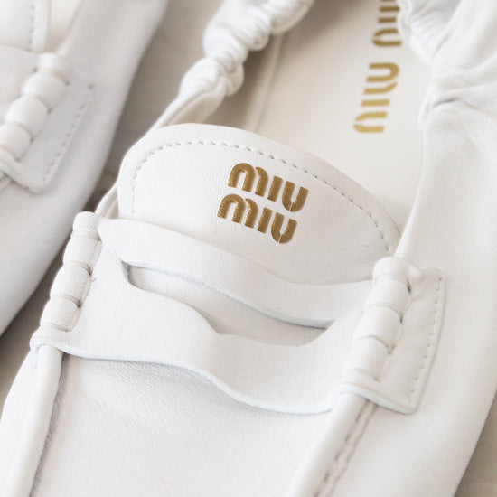 A close-up of a pair of white Miu Miu leather penny loafers with gold logo detailing on the insole and strap.