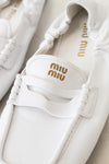 A close-up of a pair of white Miu Miu leather penny loafers with gold logo detailing on the insole and strap.