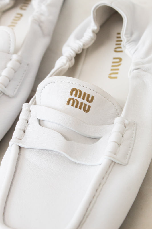 A close-up of a pair of white Miu Miu leather penny loafers with gold logo detailing on the insole and strap.