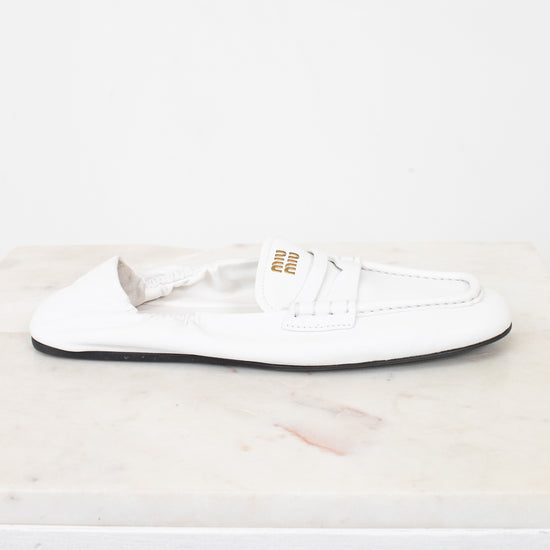 A single white leather Miu Miu loafer flat shoe with gold branding on the side, resting on a white marble surface against a white background.
