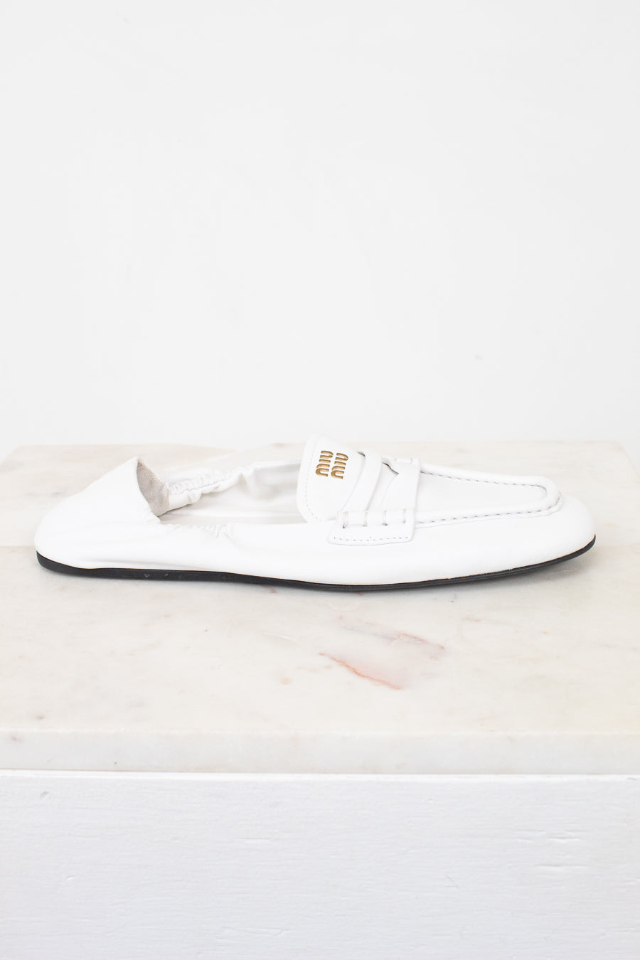 A single white leather Miu Miu loafer flat shoe with gold branding on the side, resting on a white marble surface against a white background.